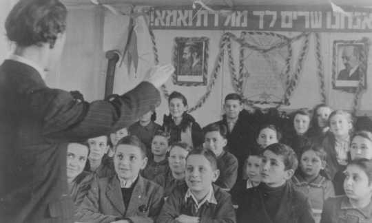 Lyrics to the Jewish national anthem and portraits of Zionist leaders hang in a classroom. [LCID: 64047]