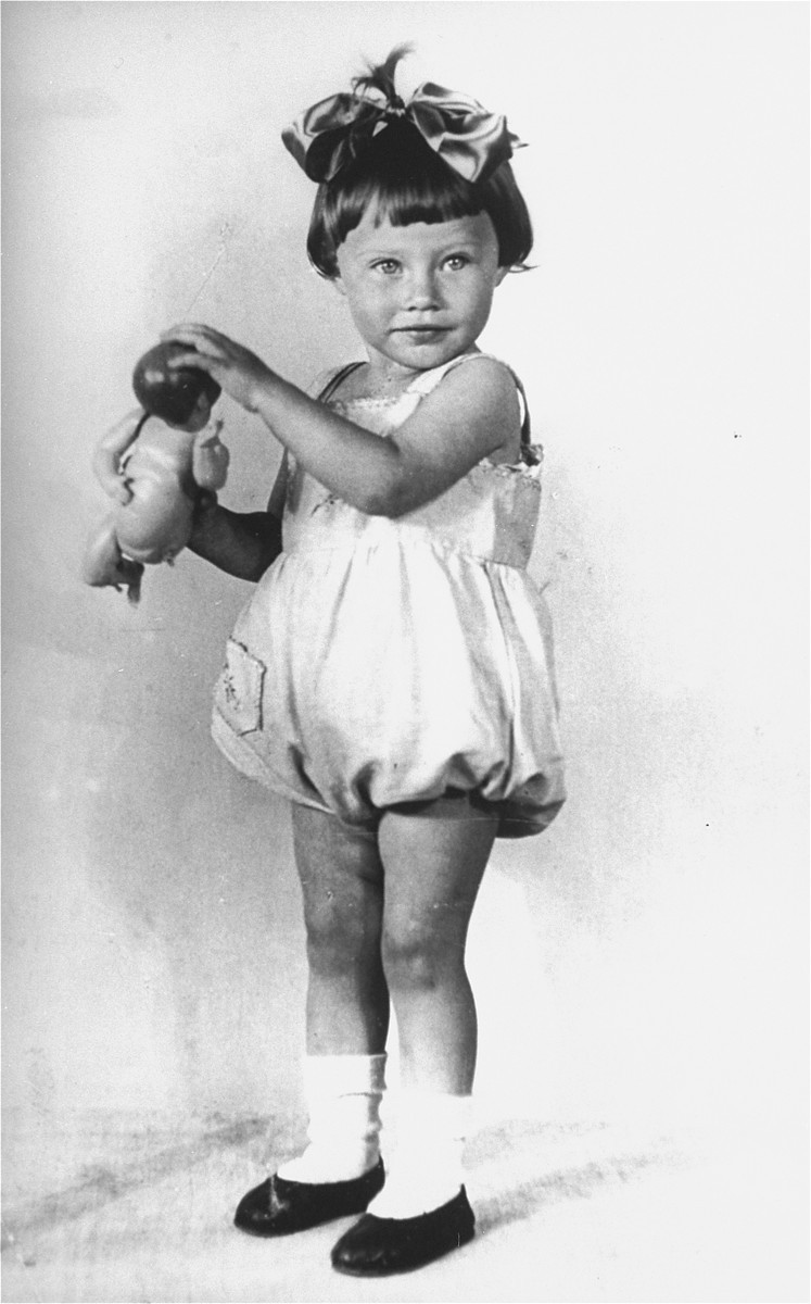 A young girl with light skin tone stands against a white background holding a doll out to the left. She is wearing a large bow in her dark brown hair and a light colored jumper, white socks, and dark shoes.