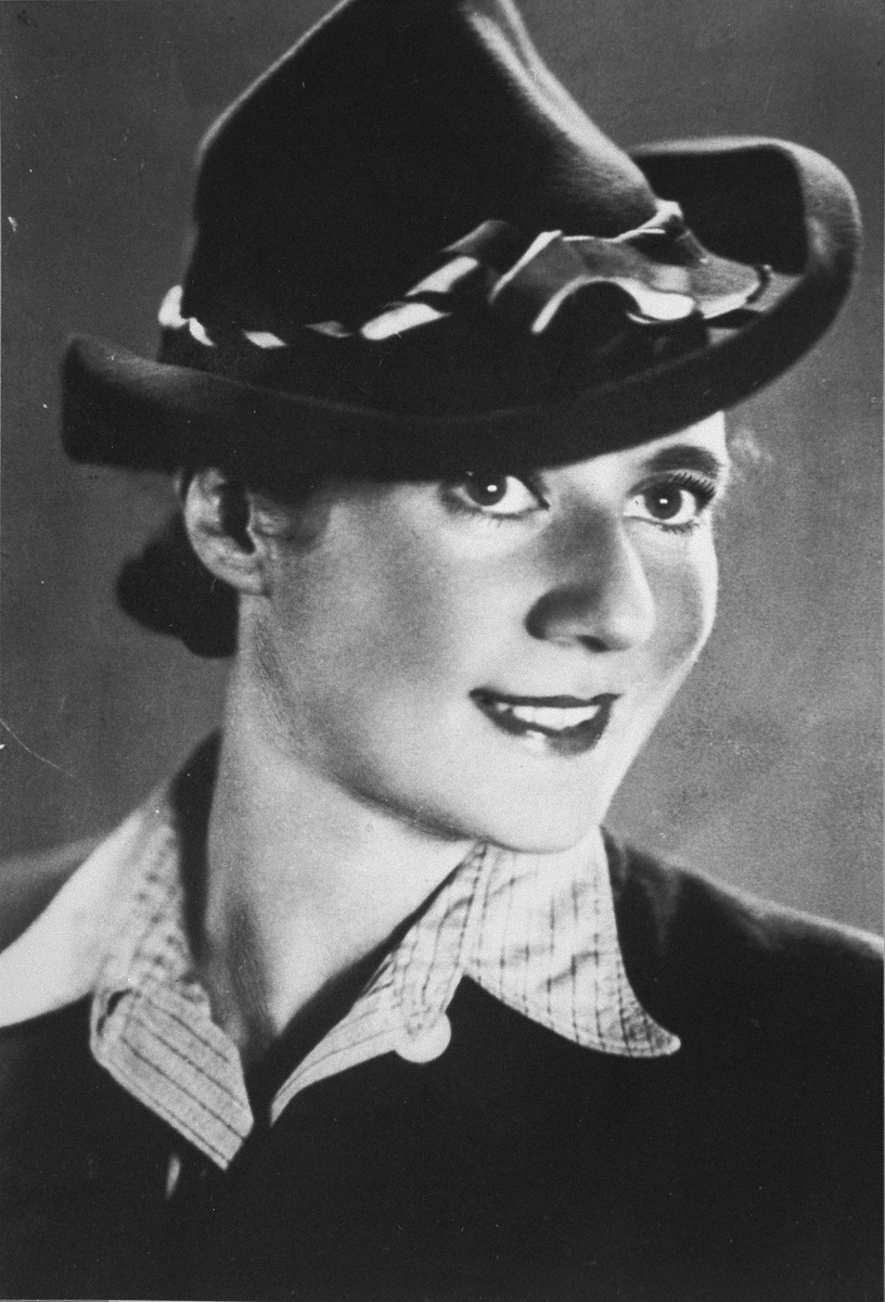 A woman with light skin tone smiles at the camera. She is wearing a collared shirt and a large fedora-type hat cocked to the side.