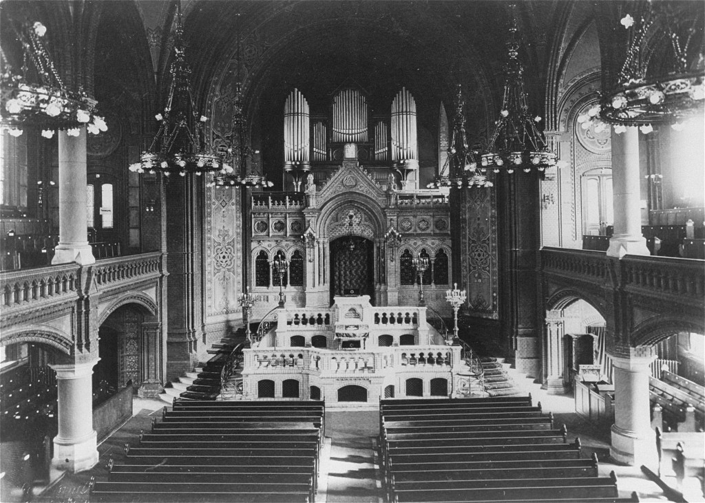 The interior view of a large, ornate, two-story synagogue with pews in the central area and women's galleries, or balconies, along the sides. At the center of the photograph sits the bimah, or synagogue platform, and the aron kodesh, or holy ark, where the Torah Scrolls are kept.
