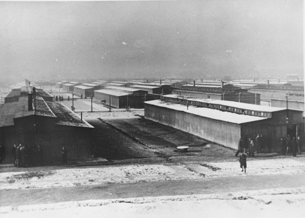 Lines of barrack buildings extend into the horizon. There is snow on the ground and on top of most of the buildings. A singular person walks through the field in front of the buildings towards a small group of people gathering near the front of one of the barracks.