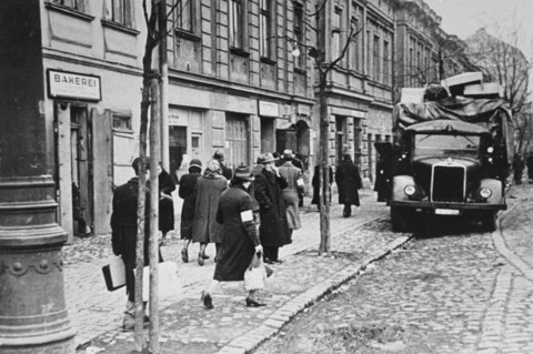 Residents of the Krakow ghetto walk past a German truck loaded with furniture confiscated from Jews. [LCID: 49002]
