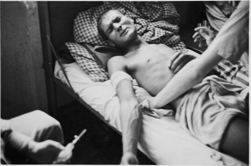 An emaciated man with light skin tone lays on a cot with a wrap around his upper right arm. The veins in his arm are pronounced and visible. He grimaces while a figure leans over the bed.