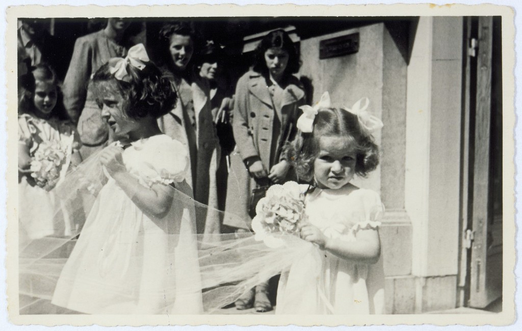 Two little girls with curly hair pulled up in updos and light skin tone stand among a group of people. They wear light colored dresses and are holding onto small bouquets and what appears to be a veil. A group of people gather in the background to watch the procession.
