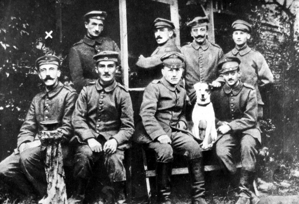 A group of eight uniformed soldiers with light skin tone arranged in two rows. The back row is standing, while the bottom row is sitting on a bench. There is a mostly white dog sitting between two soldiers in the front row.