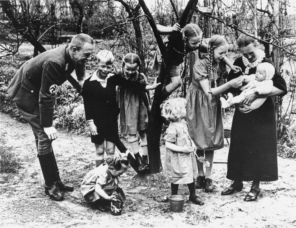 A spontaneous family portrait in a garden. There are seven children total and two adults. The presumed father wears a Nazi uniform and leans down to observe and chat with the two children to his right and the one by his feet. To the right, three children look at a small infant in the arms of the presumed mother. One of the older children has climbed a small tree to get a better look.