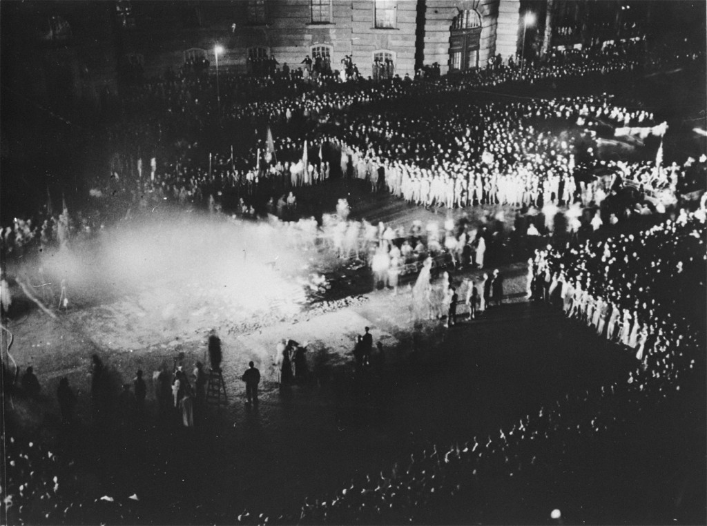 Dark black and white photograph of a city square at night, shot from above from a nearby building. A large bonfire roars in the center of the square while large groups of people gather to watch others line up and throw books into the fire.