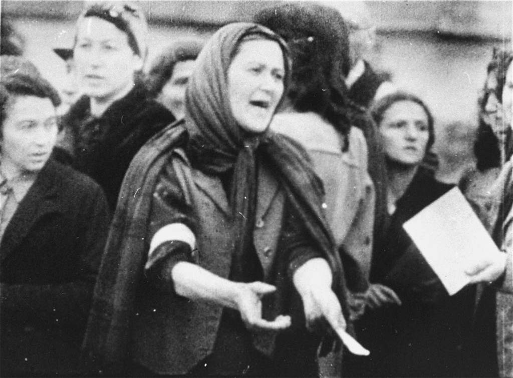 A  woman stands wearing multiple layers of clothing in a crowd. She appears to be middle-aged with light skin tone. Her hair is covered. Her hands are stretched out in front of her while she calls out to someone out of frame.