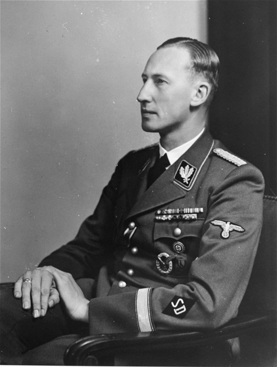 Portrait of a man sitting, dressed in a Nazi uniform. He is turned to the left with his hands folded neatly in his lap. He has short medium-dark hair and a medium-light skin tone.