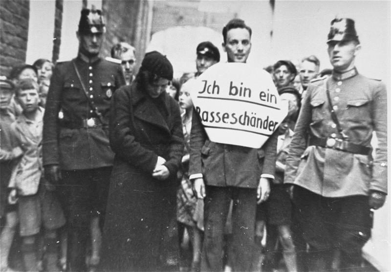 This photograph shows Julius Wolff, a young Jewish man, and Christine Neemann, his non-Jewish fiancé, standing between two police officers in Norden, Germany. Local SA men had accosted the couple and led them through the streets. The parade was meant to mock and humiliate the couple. Wolff wears a sign that reads: "I am a race defiler" ("Ich bin ein Rasseschänder"). Other photographs of this event show, and Neemann's testimony confirms, that Neemann was also forced to wear a sign. Neemann and Wolff were temporarily imprisoned in concentration camps, but were eventually released. Wolff immigrated to the United States in 1938. Norden, Germany, July 1935.