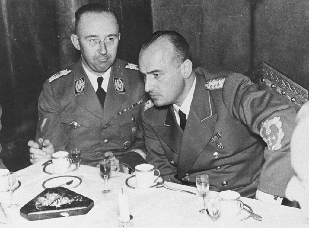 Two uniformed Nazi officials sit together at a table. The one on the right is bent over slightly with his arm resting on his knee. They appear to be drinking tea together.