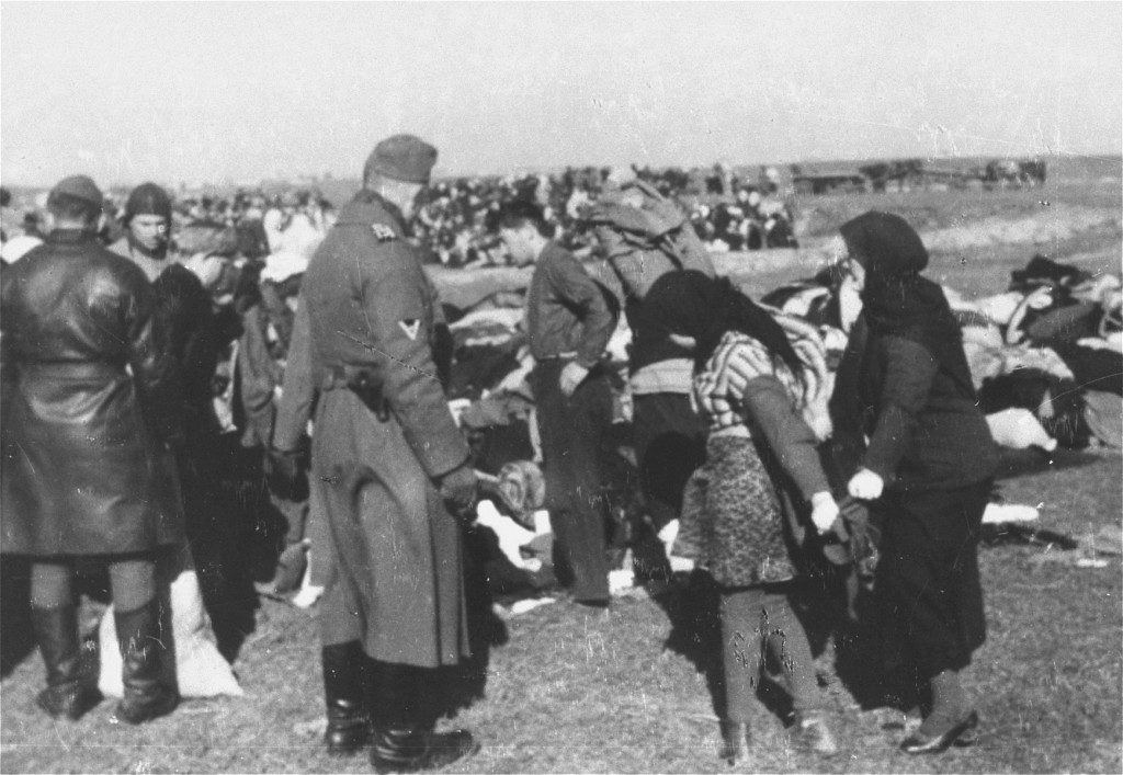 A large group of people undress in a field. There are various piles of clothing scattered around. Uniformed German soldiers observe.