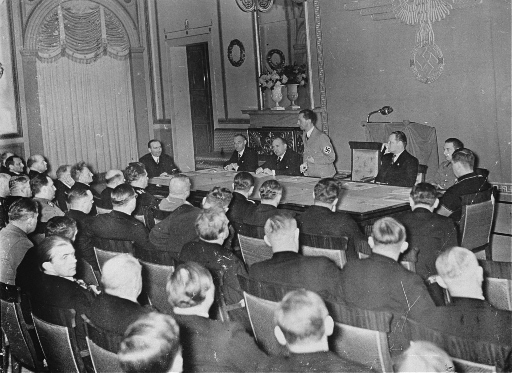 Joseph Goebbels stands behind a table at the front of a large room. Several men sit around the table and rows of men sit in chairs as part of the audience. Goebbels stands out, wearing a tan suit with a swastika armband while all the other men in the room wear dark colored clothing.