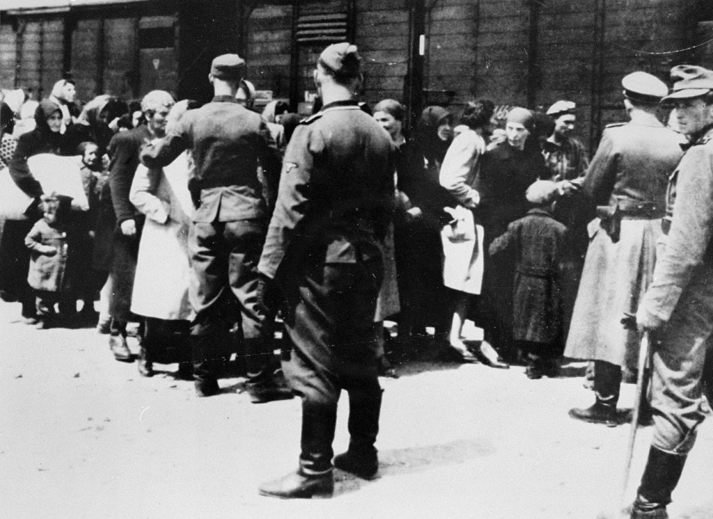 A large crowd of people stand next to a train of railcars. Several uniformed guards stand around the crowd. Only the guard with light skin tone, on the far right, has a visible face. The rest face away from the camera.