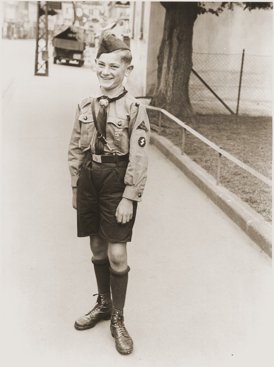 A young boy with light hair and light skin tone stands in the center of a sidewalk smiling towards the camera. He wears a full Hitler Youth uniform, complete with cover.