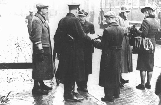 <p>Jewish police and a German soldier check documents at an entrance to the Warsaw ghetto. Warsaw, Poland, ca. 1940.</p>