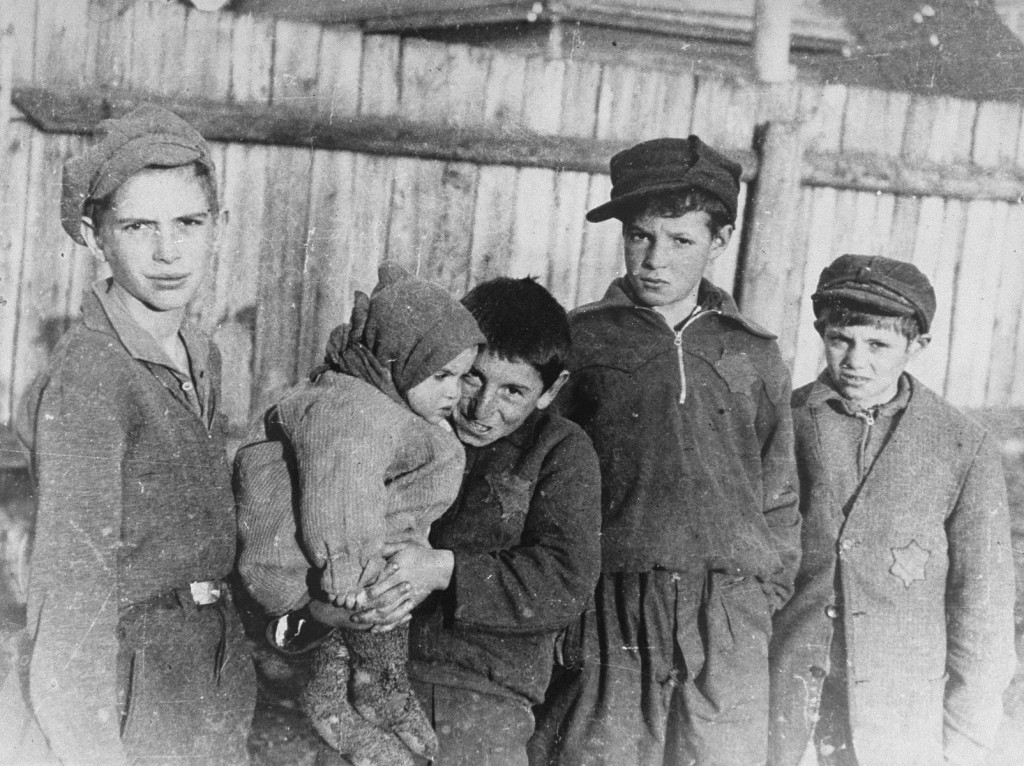 A group of five children standing in front of a fence. All of them are wearing heavy outerwear and caps. The child in the center is holding the youngest in their arms.