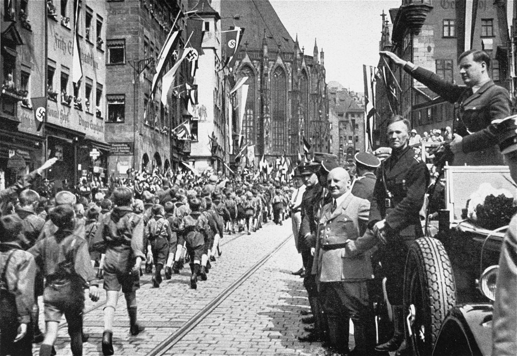 A parade of uniformed young people march down a crowded celebratory street. A group of Nazi officials stand to the right observing the parade.