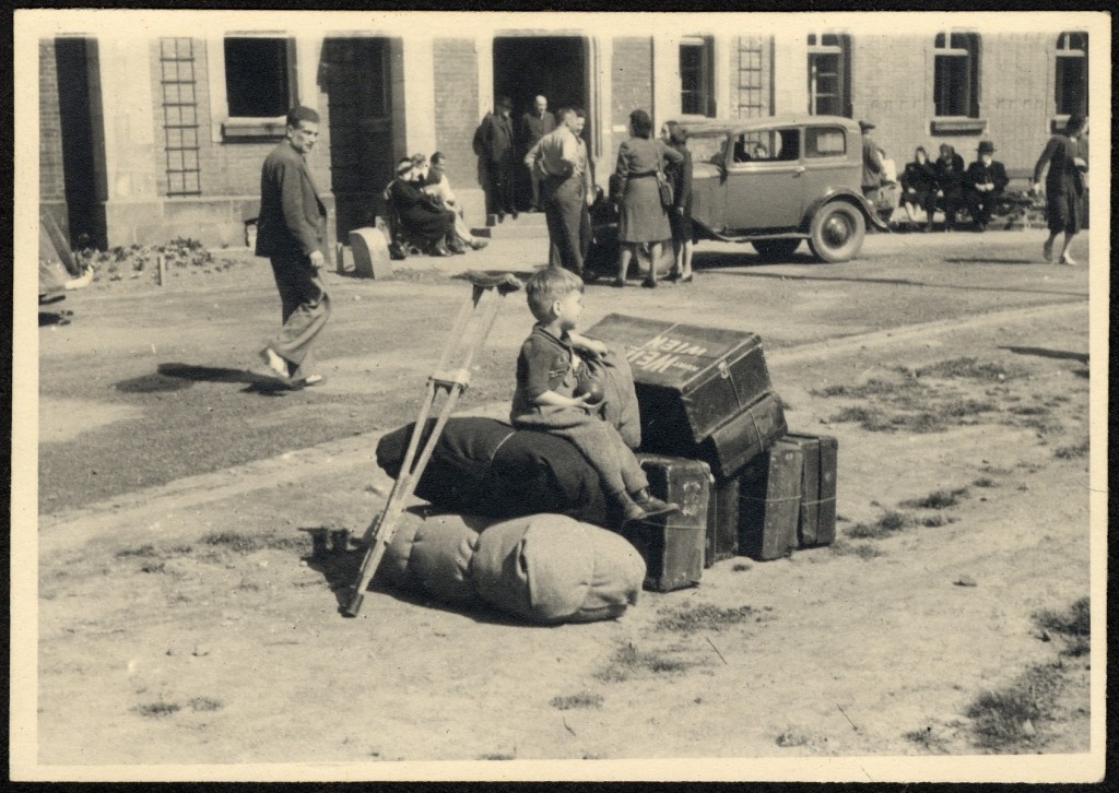 A small child sits on top of two large duffel bags with several suitcases next to him. A crutch leans on the duffel bags. People glance while walking past them on the road.
