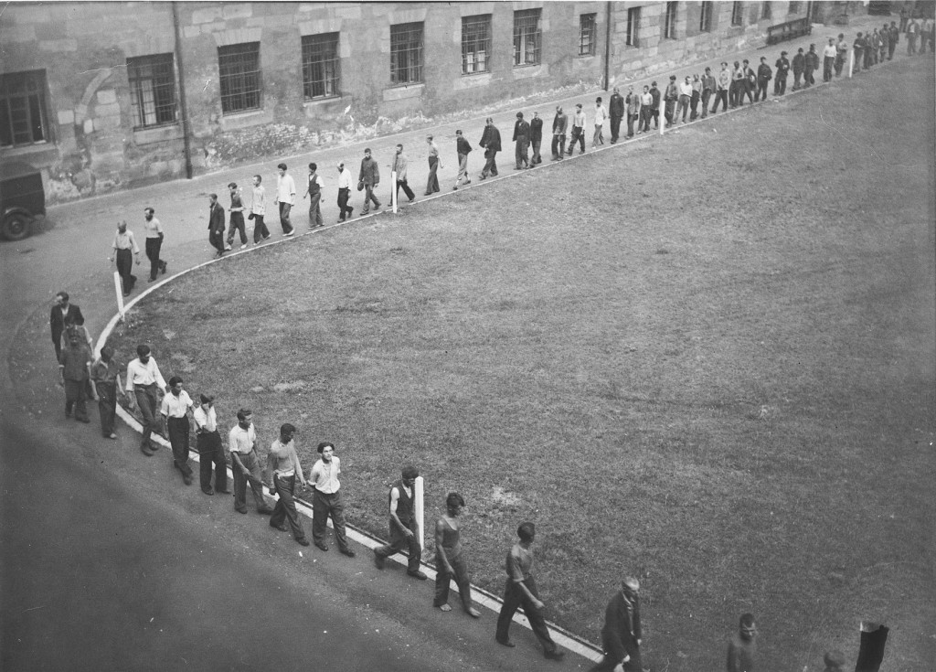 A long line of prisoners curves around a curbed green area. A large building can be seen in the background.