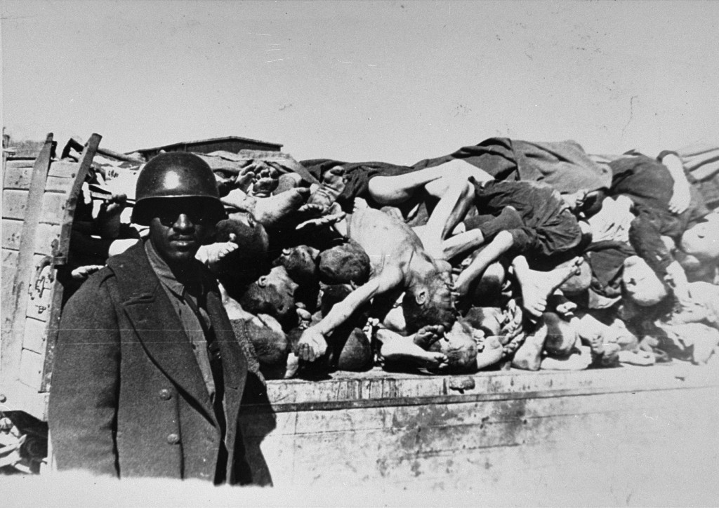 An American soldier on an inspection tour of Buchenwald poses for a photograph beside a wagon laden with corpses. [LCID: 85276]