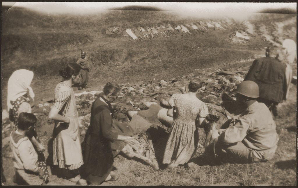 A line of people look towards a ditch filled with corpses. Uniformed soldiers observe the civilians.
