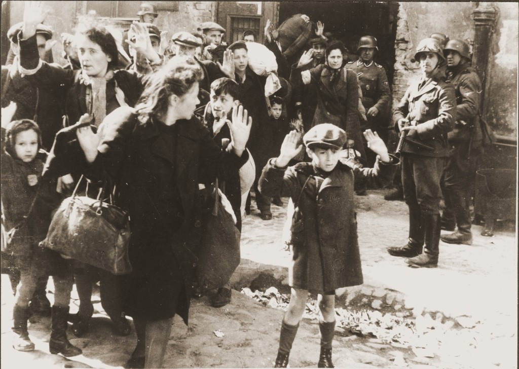 Several captured Jews, including several children, are paraded through the street with their hands up over their heads. A few German troops observe with rifles raised.