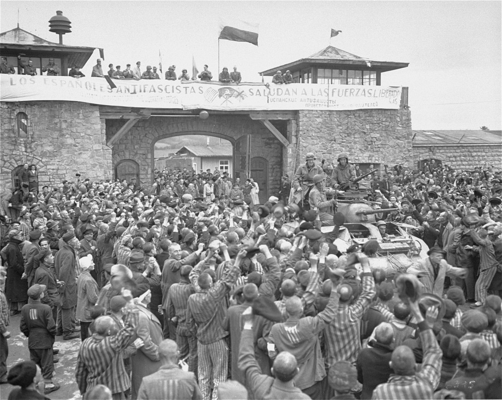 A large cheering crowd makes way for a column of soldiers and military equipment, including a tank. They are moving through a large stone gate with two lookouts on either side. There is a large banner hung across the top of the gate.