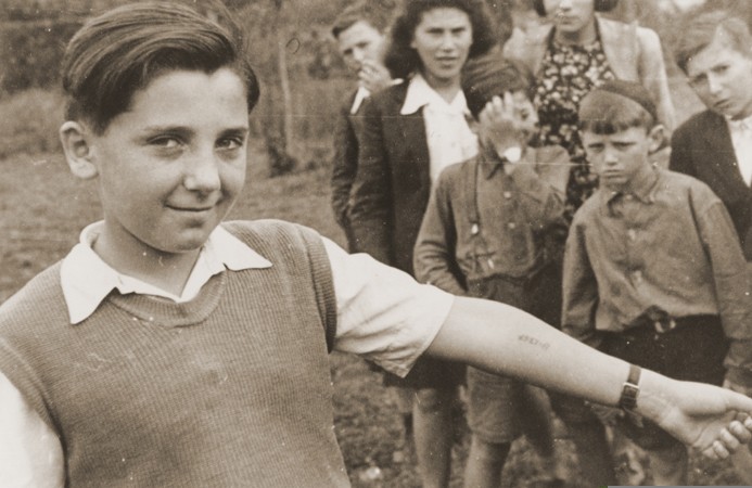 A boy displays the tattooed number on his arm to a photographer, while other children at the Neu Freimann displaced persons camp ... [LCID: 29314]