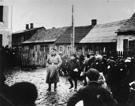 <p>A German police officer stands in a fenced-in yard among a group of Jews, who have been rounded-up in the Ciechanow ghetto. Ciechanow, Poland, 1941-1942.</p>