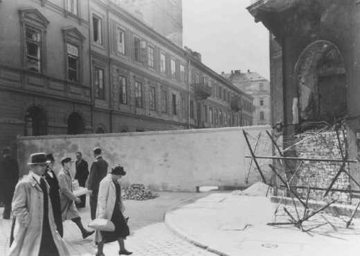 Polish civilians walk by a section of the wall that separated the Warsaw ghetto from the rest of the city. [LCID: 78390]