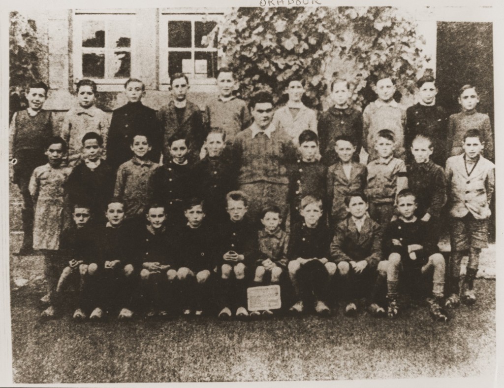 Three rows of boys line up for a school photo. A small chalkboard sits on top of one of the boy’s feet in the center of the photo. No text is visible.