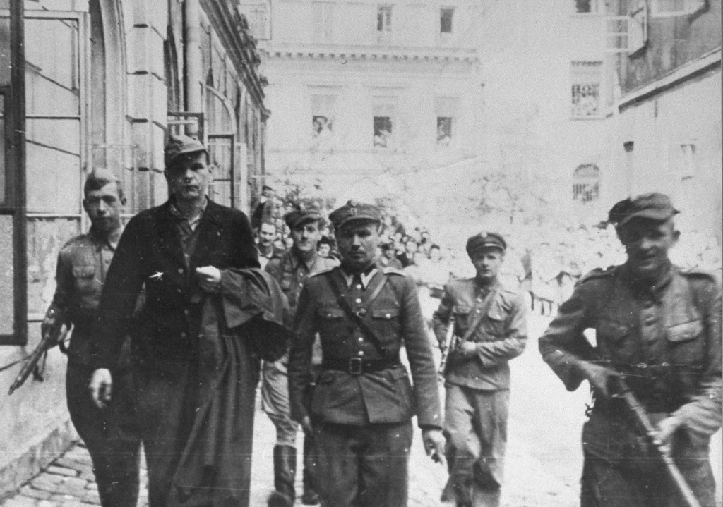 A man in military uniform is escorted down an open street by several uniformed soldiers. There appears to be a crowd behind them, watching the procession in the bright sun.