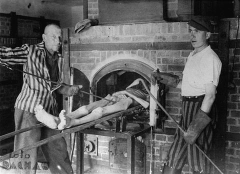Survivors of the Dachau concentration camp demonstrate the operation of the crematorium by pushing a corpse into one of the ovens. [LCID: 15026]