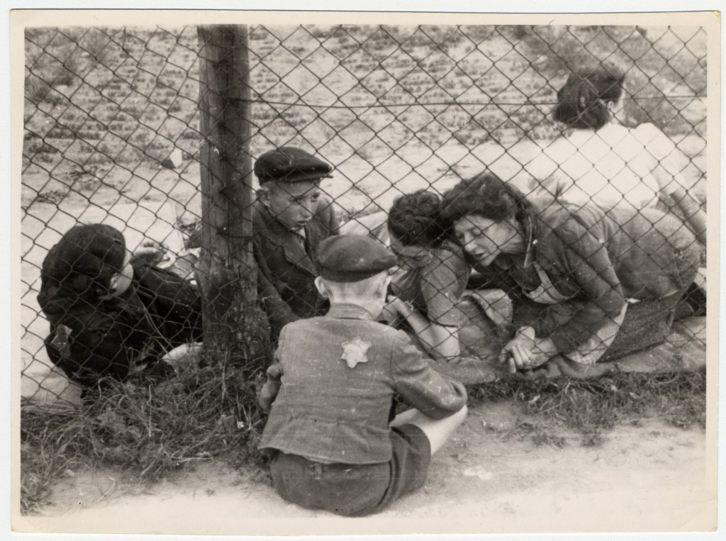 Four family members press up against a fence to say goodbye to a child on the other side of the fence. The child on the other side of the fence is sitting on the ground, facing away from the camera. A Star of David patch is pinned on the back of his coat. Another person can be seen behind the fence in the background.