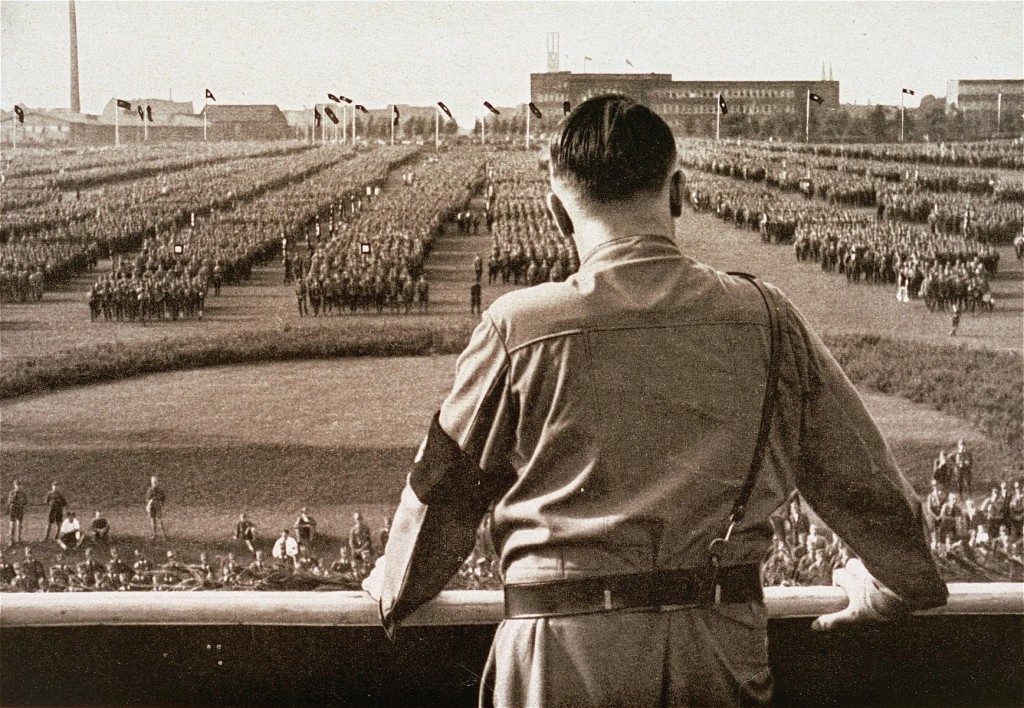 Hitler stands with his back towards the camera. In front of him is a large rally of soldiers in formation.