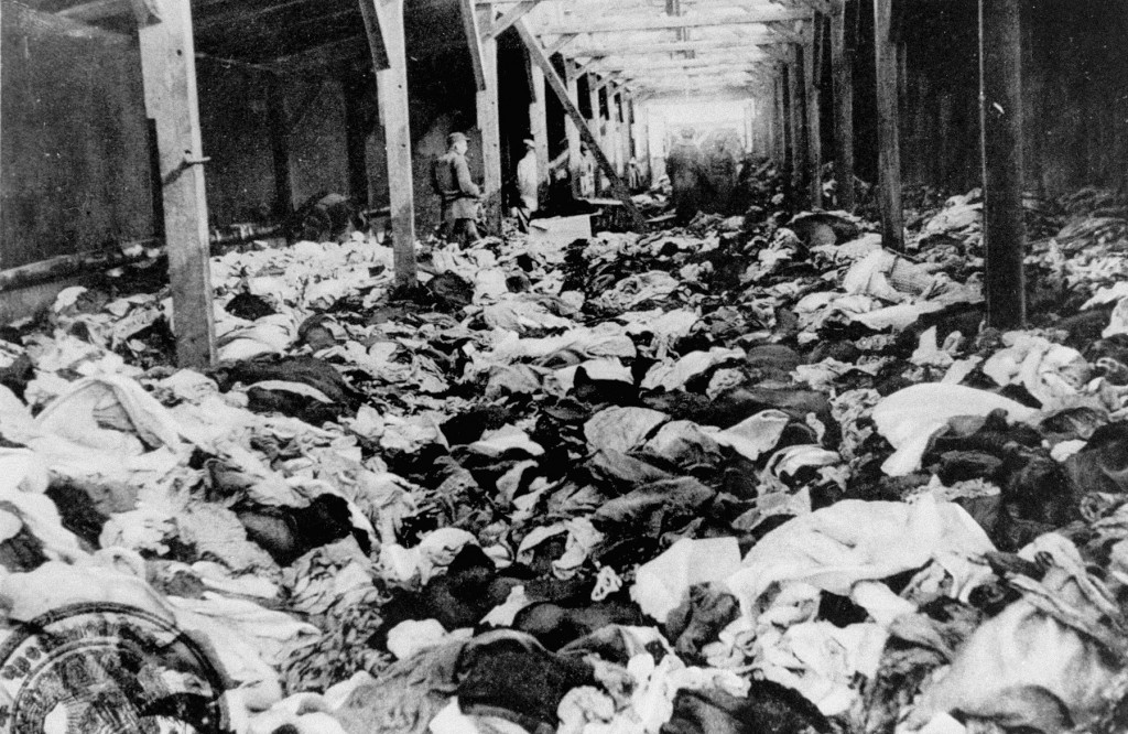 Inside of a large warehouse. The floor is littered with large piles of clothing of every shape, size, and color. A few uniformed soldiers can be seen in the background, walking amongst the clothing.