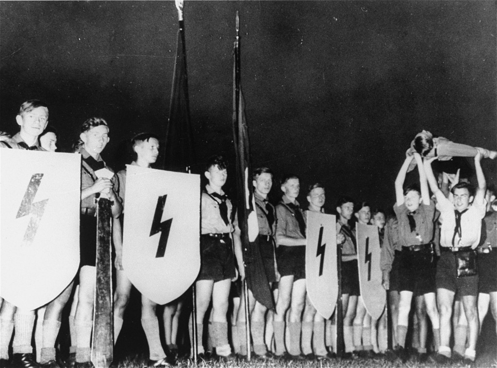 Dozens of young boys in Hilter Youth uniforms: dark shorts, light short-sleeved shirts and dark handkerchiefs stand in rows. At the front, two boys hold flags, four boys hold handmade shields with lightning bolts. A group of boys hold another on his back over their heads and parade him in front of the larger group.