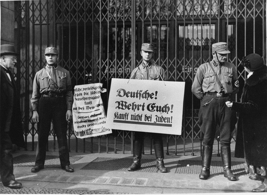 Three uniformed Storm Troopers with light skin tone stand in front of a closed shop. The soldier in the center holds a boycott sign in German, while another sign hangs from the gates covering the shop entrance. The soldier to the right appears to be speaking to a feminine figure with light skin tone in dark clothes.