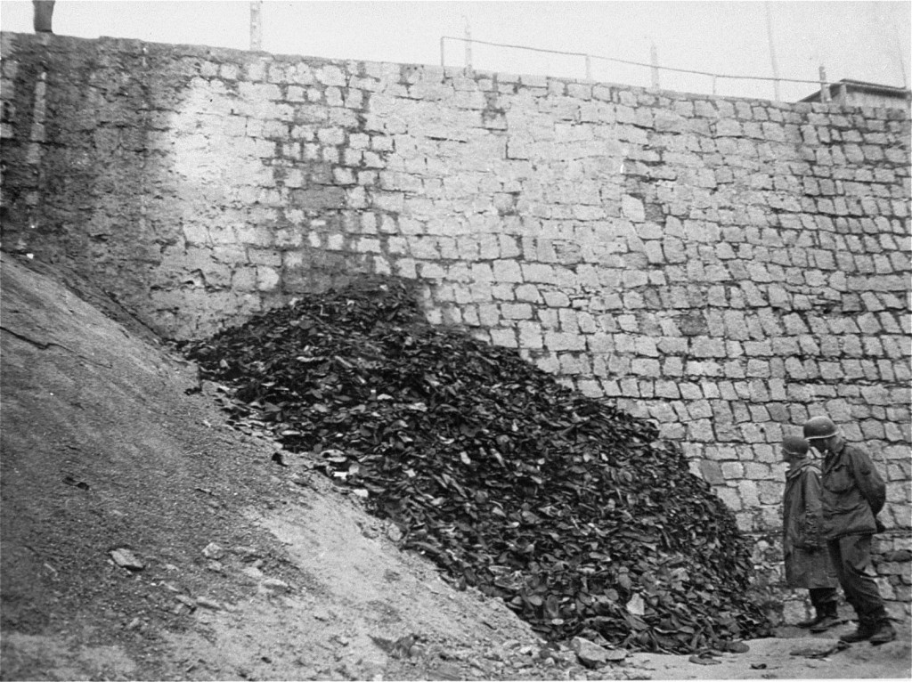 A monumental pile of leather shoes has been built against a large rock wall. The pile towers above two uniformed soldiers observing on the right.