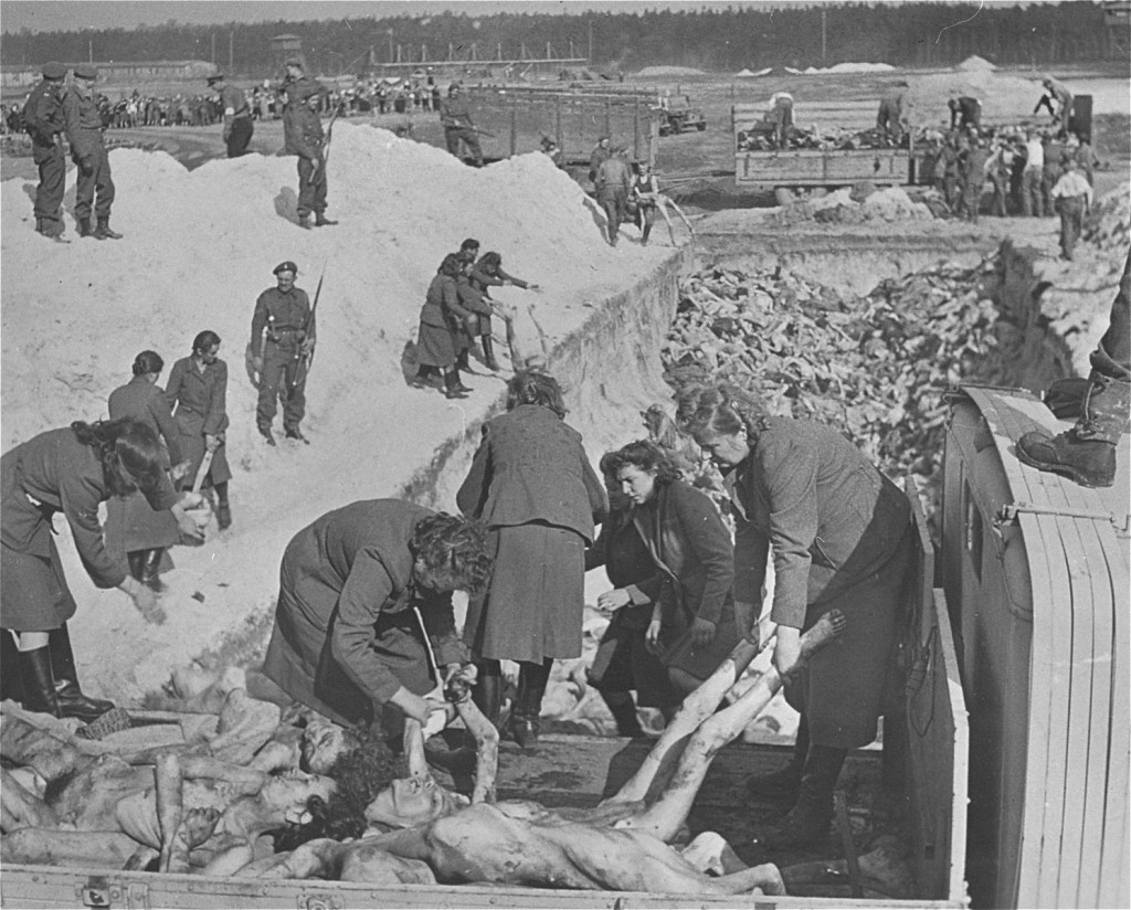 A uniformed soldier stands at the edge of a large burial pit. Groups of light-skinned women in dark clothing carry emaciated bodies by the hands and feet into the mass grave.