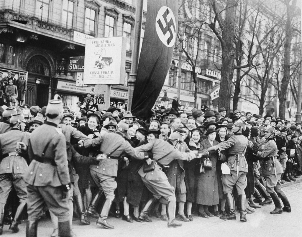 A large group of people stand along the side of a city road. Several individuals push against a line of uniformed soldiers to try and push closer, spilling out into the road.
