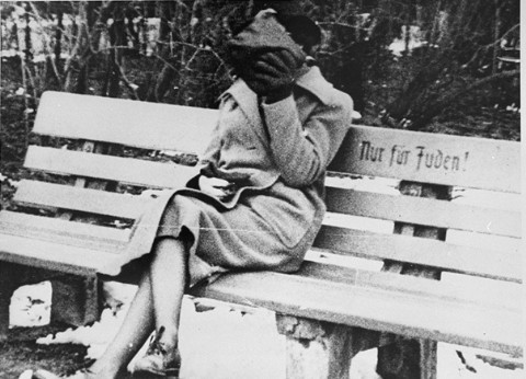 A woman covering her face is seated on a park bench that bears the inscription Nur für Juden! (Only for Jews)