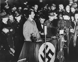 Goebbels speaks at a swastika draped table. Microphones are arranged in front of him while reporters, uniformed servicemen and other individuals gather around.