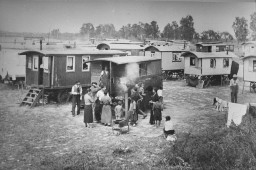 A large open field with several parked wagons. People of all ages gather around one wagon with a wood burning stove and laundry hanging on a drying line.