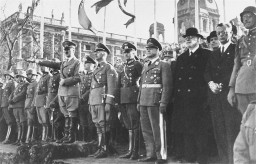 Adolf Hitler stands at the head of a large group of uniformed soldiers and officials. He raises his arm in a Nazi salute towards a parade occurring out of frame.