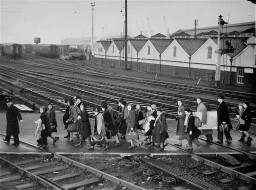 A group of children walk together over a large grouping of train tracks. They are all wearing coats and carrying luggage. A few adults can be seen at either end of the group.
