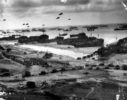La playa de Normandía como se veía después del Día D. Embarcaciones que llegan a la playa descargan tropas y suministros transferidos de transportes costa afuera. Globos de barrera se sostienen en el aire para disuadir a los aviones alemanes. Sin fecha.