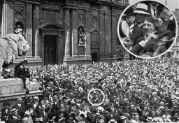 A large crowd gathers in a large plaza celebrating the outbreak of war in 1914. The face of Adolf Hitler is enlarged to show his presence in the crowd.