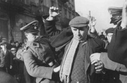 German personnel on Grzybowska Street arrest and search Jewish men who supposedly hid weapons prior to the German occupation of Warsaw.  Warsaw, Poland, October-December 1939.
This is one of a series of photos taken by Arthur Grimm, an SS propaganda company photographer, documenting the investigative work of the Sicherheitsdienst (SD) in occupied Warsaw for the Berliner Illustrierte Zeitung. Although only some of the photos were published, it is likely that the incidents depicted in the BIZ were staged for propaganda purposes, even despite the fact that the SD did indeed conduct searches for weapons in Polish cities during the first months of the German occupation. According to Grimm's report, a Polish man informed the SD that Jews had hidden weapons. Several (4 to 6) men were then arrested and supposedly confessed to burying weapons in the grave of a Polish soldier. Four Jews were subsequently brought to the site of the grave and forced to dig up the weapons. Afterwards, their beards were publicly shorn and the "suspects" were made to stand trial before a German judge.